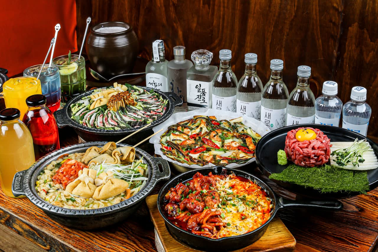 밀푀유나베와 전골, 육회를 가운데 두고 둘러놓은 한 상, 뒤쪽에 ‘새벽’ 라벨이 붙은 수제 막걸리 병들이 서 있다