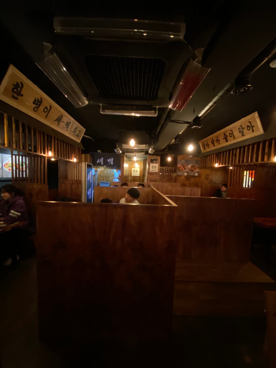 어둑한 조명 아래 나무 칸막이로 나뉜 좌석들과, 천장에 걸린 손글씨 현수막 — 안쪽에 손님들이 앉아 있다
